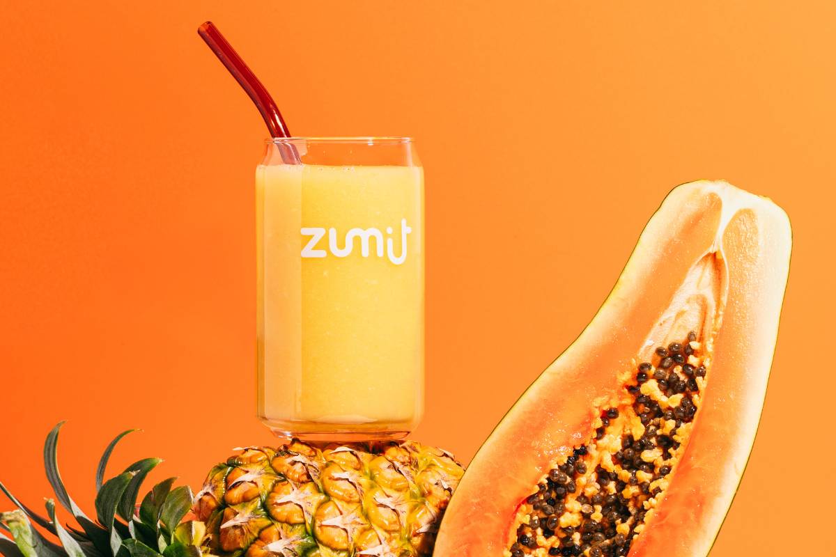 Smoothie Zumit Caribbean Passion con mango, papaya y piña apoyado sobre una piña y una papaya